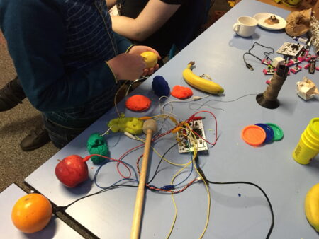 Lumps of brightly coloured Play Doh with wires inserted into it. Sitting alongside the Play Doh are an orange,an apple and a banana,all have wires inserted.All the wires are connected to a small circuit board.