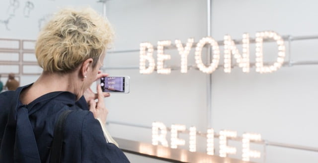 The words " Beyond Belief " written in large white writing on a wall. In the foreground is a person taking a photo with a smart phone.