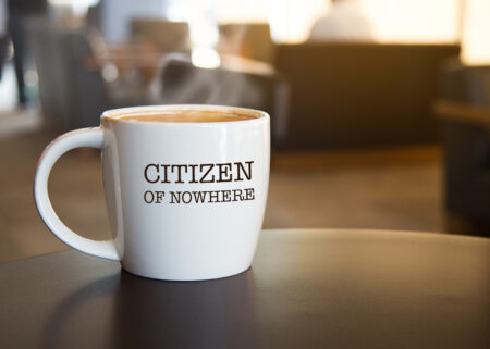 A large white mug filled with steaming coffee sits on a round,brown table. The words "Citizen of Nowhere" is written on the side of the mug in black, capital letters.