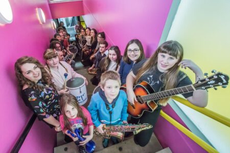 Group of young people standing in a bright pink stairwell, each holding a different musical instrument.