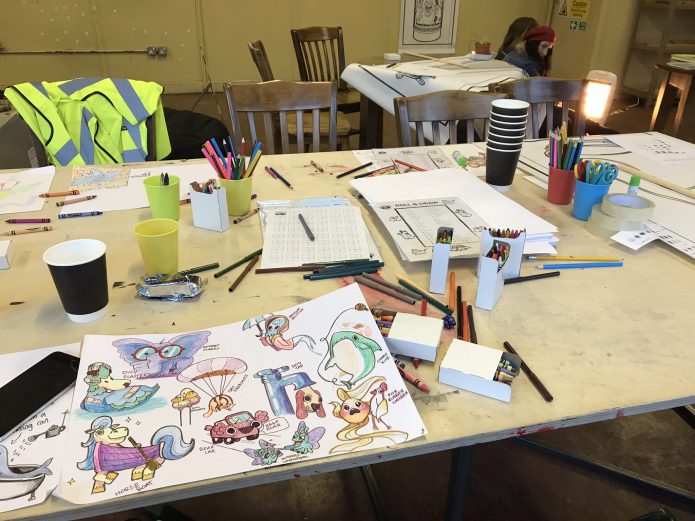 A large white table with various art materials eg, coloured pencils, pens,paper and pairs of scissors. There is also a piece of paper with various illustrated character designs.