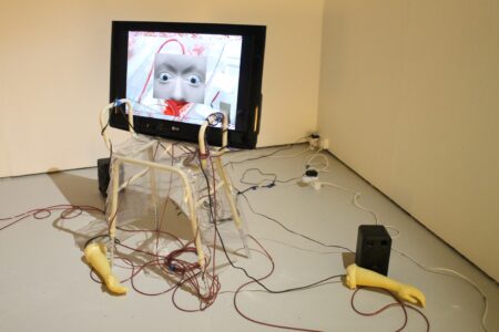 In the corner of a white room sits a black monitor screen which is sitting on a walking frame. The image on the screen is of a wide eyed robots face. Coming out of the screen are wires which wrap around the walking frame and onto the floor. These wire are attached to two plastic arms which are lying on the floor.