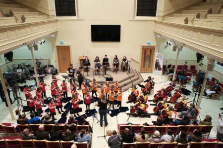 An aerial view of a large group of school children playing violins and cellos to a small audience of people.