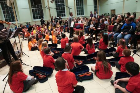 In a large Hall, a person talks to a seated adult audience. Sitting on the floor in front of the audience are a group of school children with their violins and cellos.