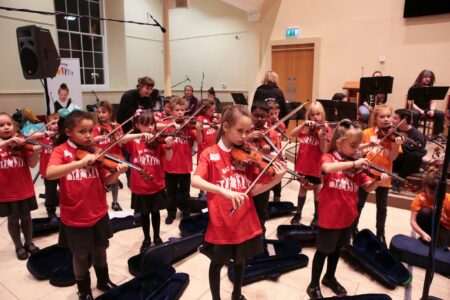 A group of school children playing violins in a large hall.