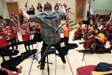 A person standing in a large room,possibly a concert hall, is conducting a group of children playing violins and cellos.