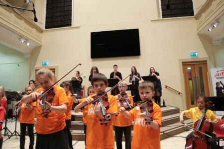 A group of school children,wearing orange t-shirts,playing violins. In the background are 5 people,dressed in black, playing woodwind instruments.