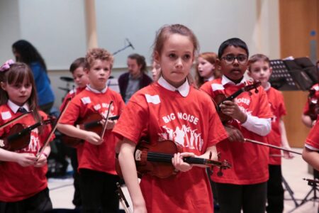 A group of school children wearing red t-shirts, holding violins.