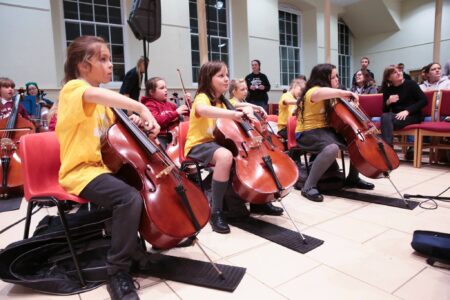 Three school children sitting in the middle of a large room, playing cellos'.