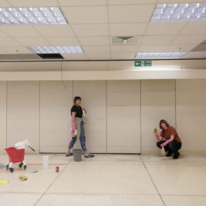 Two people in a white room, one standing the other crouching, with various cleaning materials.