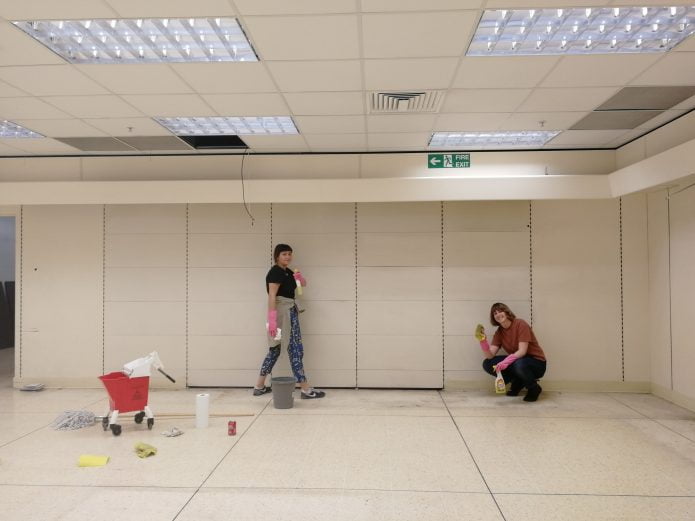 Two people in a white room, one standing the other crouching, with various cleaning materials.