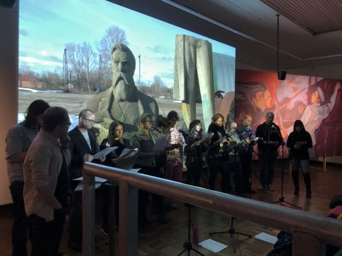 A group of people with microphones standing in front of a screen which is showing an image of a statue. The statue is in two halves and is situated in what looks like an empty carpark.