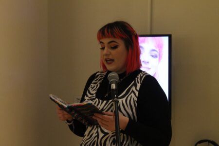 Artist,Megan Goldie,reading from a book into a microphone.