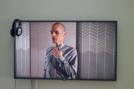 A wall mounted television,on a white wall. On the screen is a bald headed person wearing a grey shirt and tie,standing in front of patterned wallpaper.On the corner of the television are a pair of headphones.