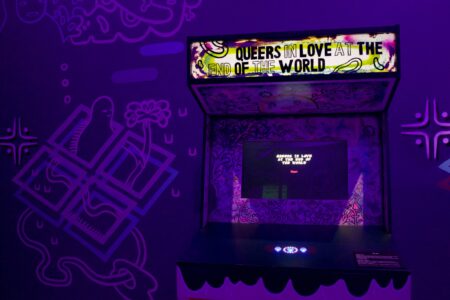 An arcade game called Queers in Love at the end of the world, standing in a dark,purple coloured room.