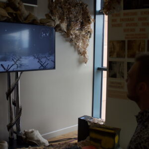 A person sitting in a room which has various photographs and writings on the walls, playing a nondescript video game.