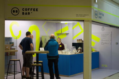 Three people standing in a shop kiosk. The kiosk is off white in colour with large yellow writing,spelling out the word "Coffee". Standing behind the blue counter is another person, engaging in conversation with the other people in the kiosk.