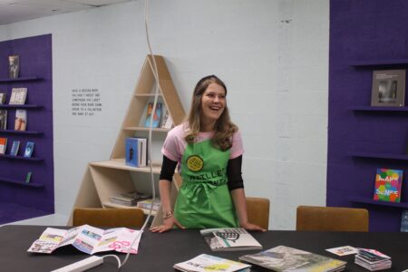 A person,wearing a pink t shirt and a green apron stands behind a desk which has booklets of various sizes on top of it.Behind this person are three bookcases with books on them. The bookcase in the middle is in triangular in shape, the bookshelves either side of this are both purple in colour.