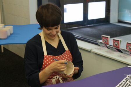 A person wearing a red apron with with white spots is holding a large cup/mug in one hand and a pencil in the other.