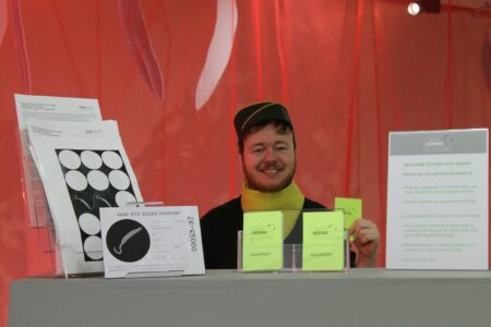 in front of a red curtain is a person wearing a black and yellow air hostess uniform, behind a large,grey counter. On top of the counter are various 'brochures' with the the words " Para-Site-Seeing Passport" written on most of these 'brochures'.
