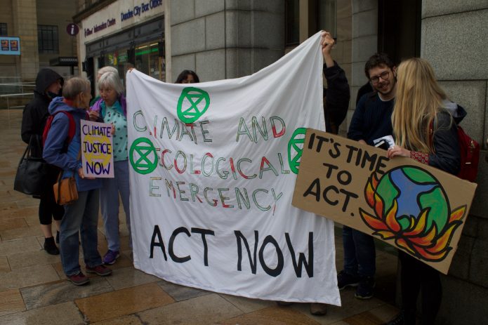 Extinction Rebellion Protesters holding a large white banner which reads," Climate and Ecological Emergency ACT NOW".