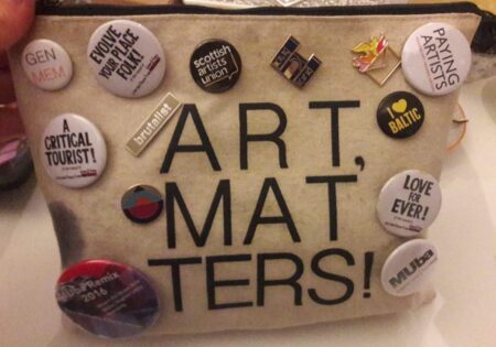 A small,off white,felt pencil case. Written in large black letters are the words," Art,Matters!". Dotted around the pencil case are various pin badges.