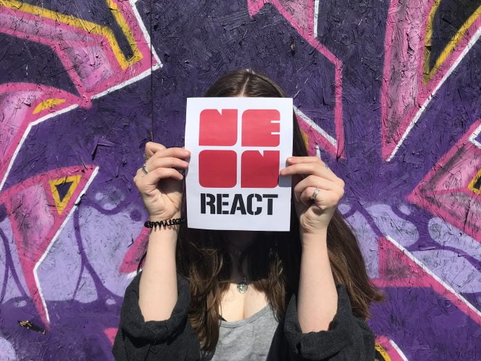 Photograph of a person standing in front of a graffiti board holding the NEoN REACT logo in front of their face.