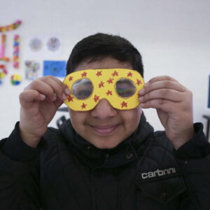 Young boy wearing a black jacket, holding up yellow fractal googles in front of his face and smiling. The googles are yellow and have been decorated with red stars.