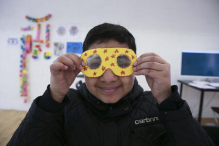 Young boy wearing a black jacket, holding up yellow fractal googles in front of his face and smiling. The googles are yellow and have been decorated with red stars.