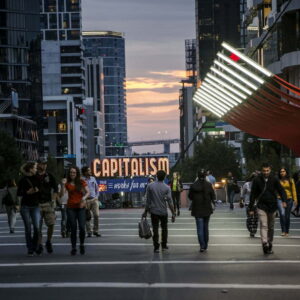 A busy pedestrian crossing, in the background are the words " CAPITALISM" written in large orange lights.