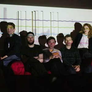 The five members of the band Field Music, sitting on a red sofa in a dark room. They are sitting in front of a white projected image with brightly coloured wavy lines.
