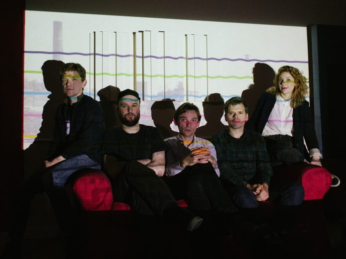 The five members of the band Field Music, sitting on a red sofa in a dark room. They are sitting in front of a white projected image with brightly coloured wavy lines.