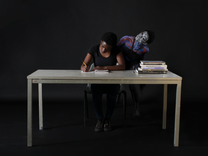 Person sitting writing at a desk whilst a person in African tribal body paint looks on.