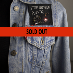 A blue denim jacket with a Stop Buying Plastic patch sewn on.Superimposed over the image are the words "Sold Out" written in black.