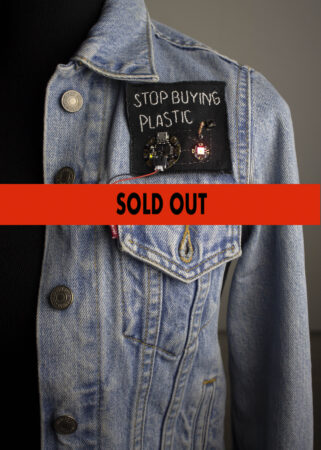 A blue denim jacket with a Stop Buying Plastic patch sewn on.Superimposed over the image are the words "Sold Out" written in black.