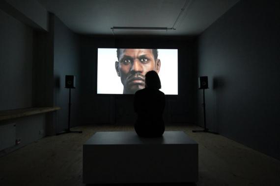 Person sitting in a small dark room watching an image of another person.