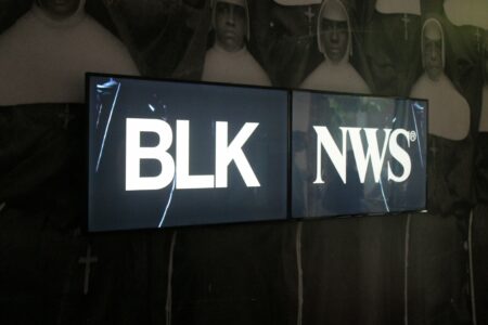 Two monitor screens standing in front of a large black and white image of a group of nuns. On the left screen are the letters BLK in white on a black background, the screen on the right has the letters NWS also in white writing on a black background.
