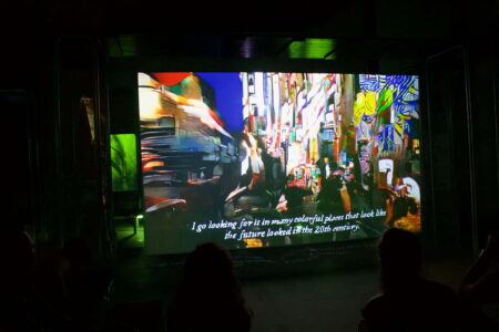 People in a dark room watching bright coloured, abstract images on a cinema screen. At the bottom of the screen are the words " I go looking for it in many colourful places that look like the future looked in the 20th century".
