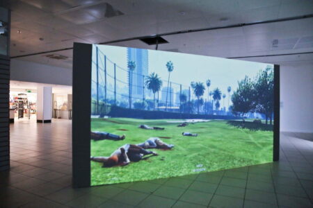 A large screen with a computer generated image of a park which has dead people lying on the grass.