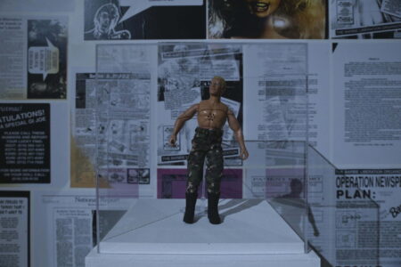 An Action man figure wearing camouflaged trousers standing in a clear perspex cube.