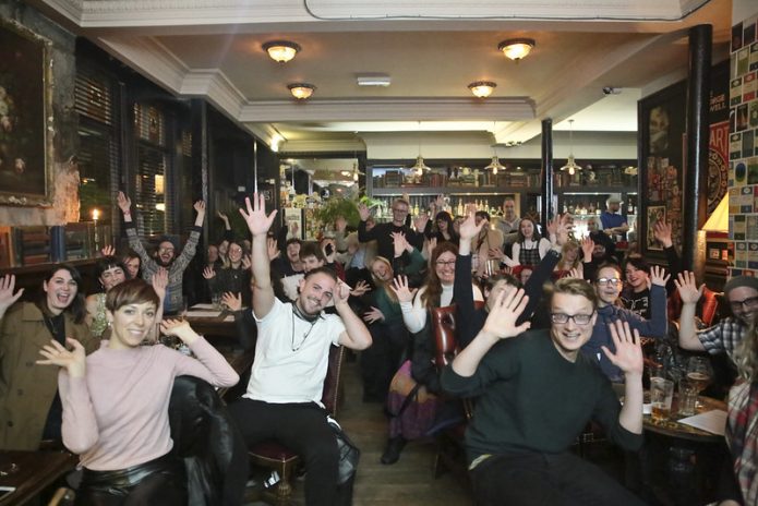 A group of approximately 50 people sitting in a bar/ restaurant,waving towards the camera.