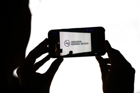 A person holding a smart phone which has the Abandon Normal Devices logo on the screen.