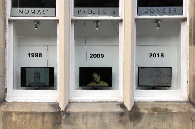 Three televisions in three shop windows. The tv on the left has the date 1998 written on the window above the tv. The tv in the middle has the date 2009 written above it. The tv on the right has the date 2018 written above it.