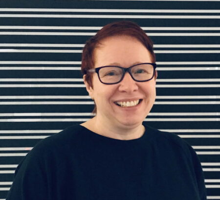 NEoN Director Donna Holford-Lovell. Donna is standing in front of a black and white, horizontally striped, background, and is smiling.