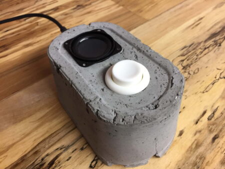 A computer mouse made from grey concrete. On top of the mouse is a small black speaker and a round white button.