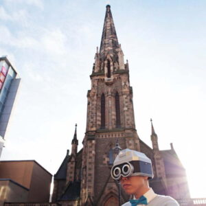 Eric Sui dressed as his alter ego Touchy. He is wearing a white shirt with a blue bow tie. On his head is a silver robot type helmet with black goggles over his eyes. he is standing in front of the Mecca bingo building in Dundee.