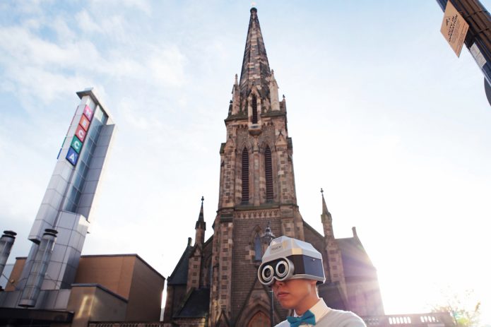 Eric Sui dressed as his alter ego Touchy. He is wearing a white shirt with a blue bow tie. On his head is a silver robot type helmet with black goggles over his eyes. he is standing in front of the Mecca bingo building in Dundee.