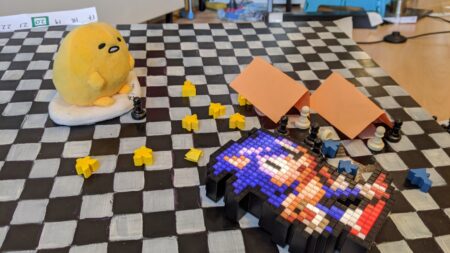 A black and white chess board type type table upon which is a yellow egg shaped plush toy, 8 black and white chess pawns, a few yellow and blue small indistinct shapes and a Sonic the Hedghog made from lego type bricks.