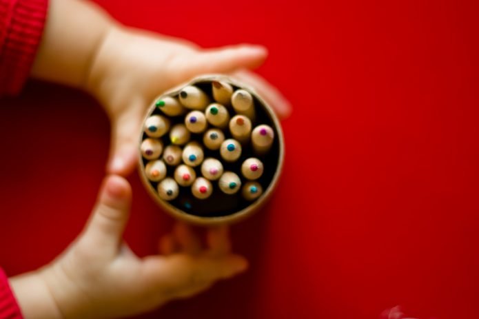 Young child hands grasping a pot of pencils on a dark red background.