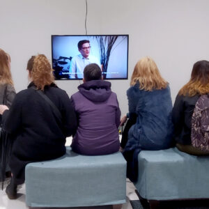 A group of Level two DJCAD students sit on grey cubes in a white room, watching a video artwork on a small screen. The video artwork is Utopia Generator by artist Julia Schicker, and the screen shows a white person in glasses in mid-sentence.